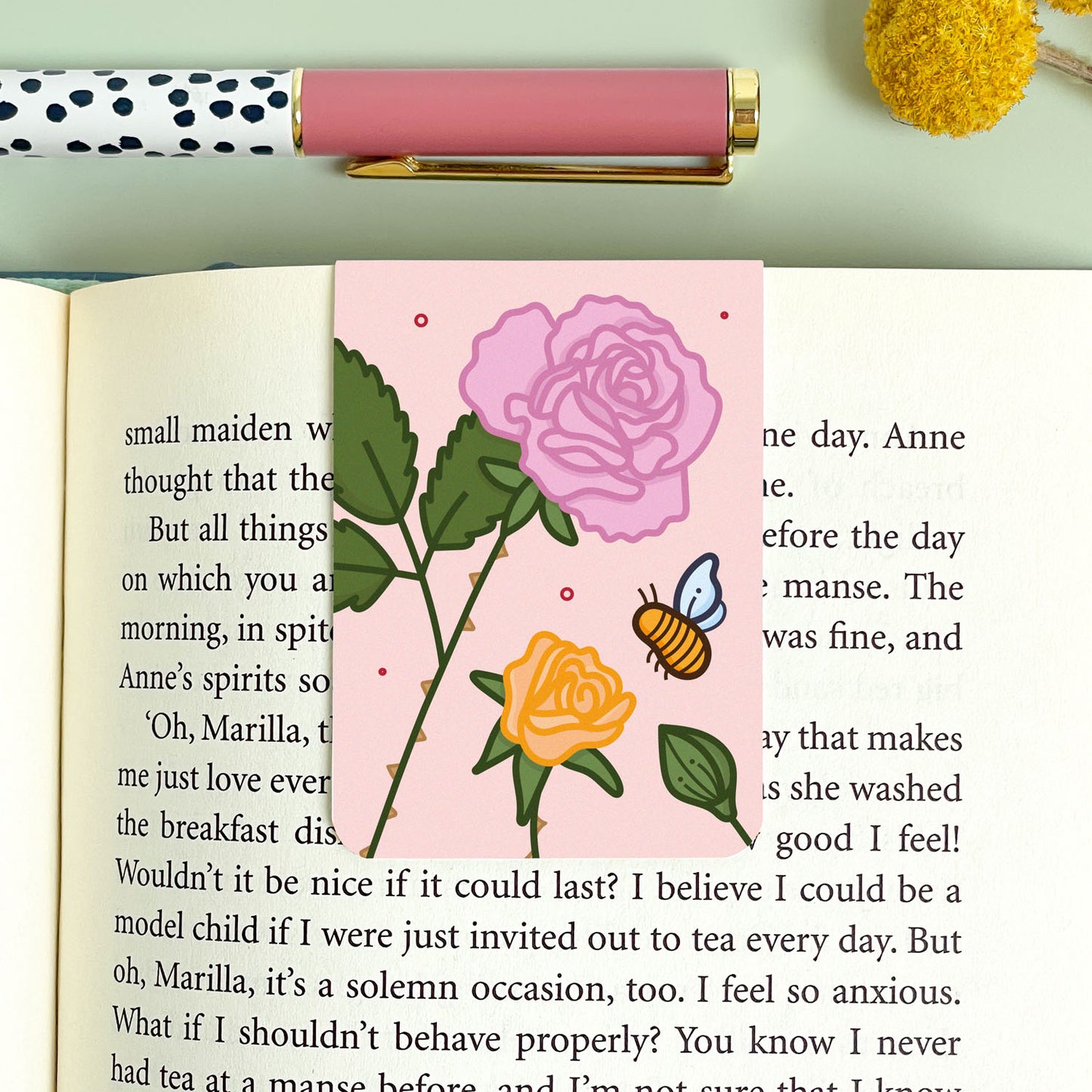 Floral rose bookmark with a bee on an open book, next to a polka dot pen.