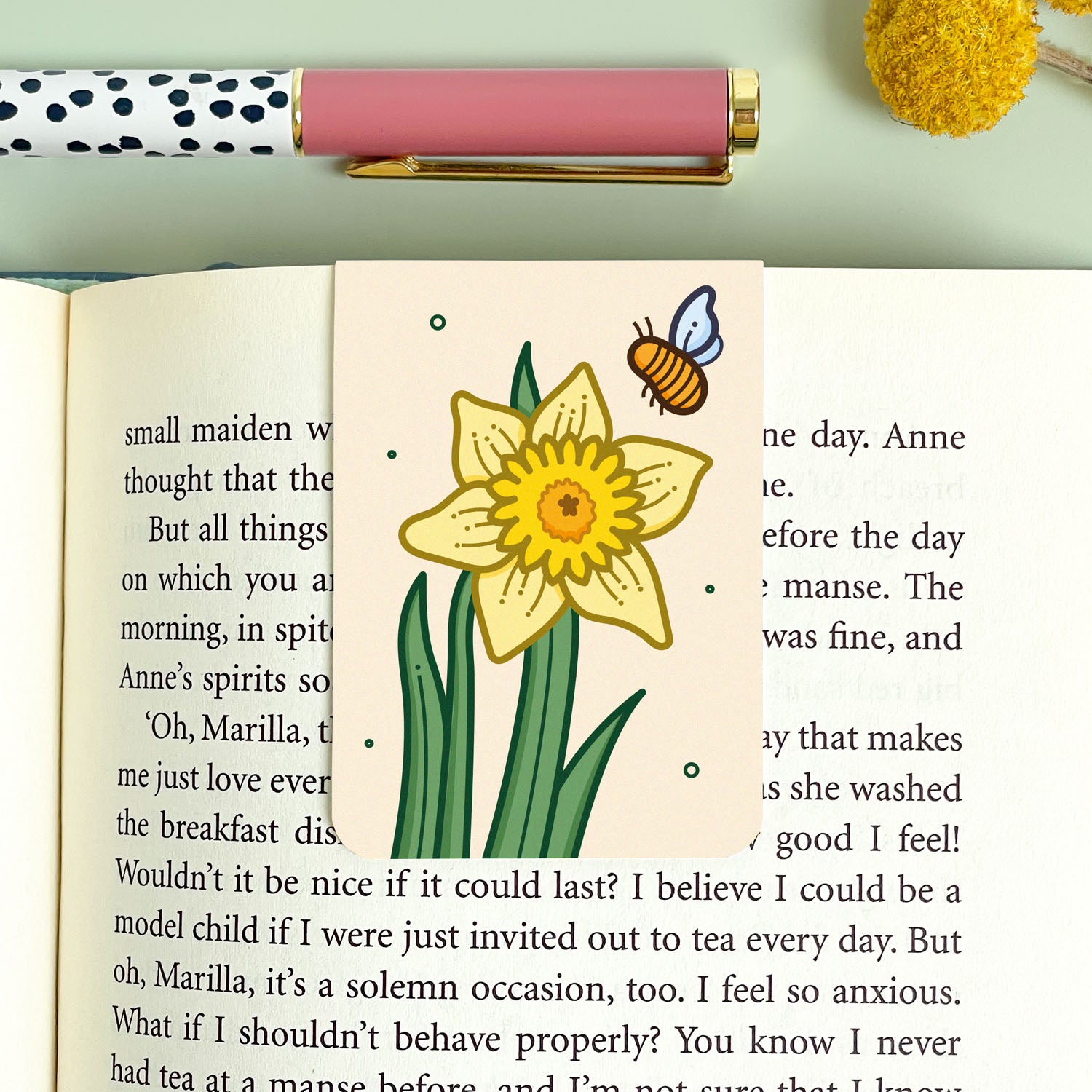 Bookmark with a yellow daffodil flower illustration and a bee, next to a pink pen on a light green background.