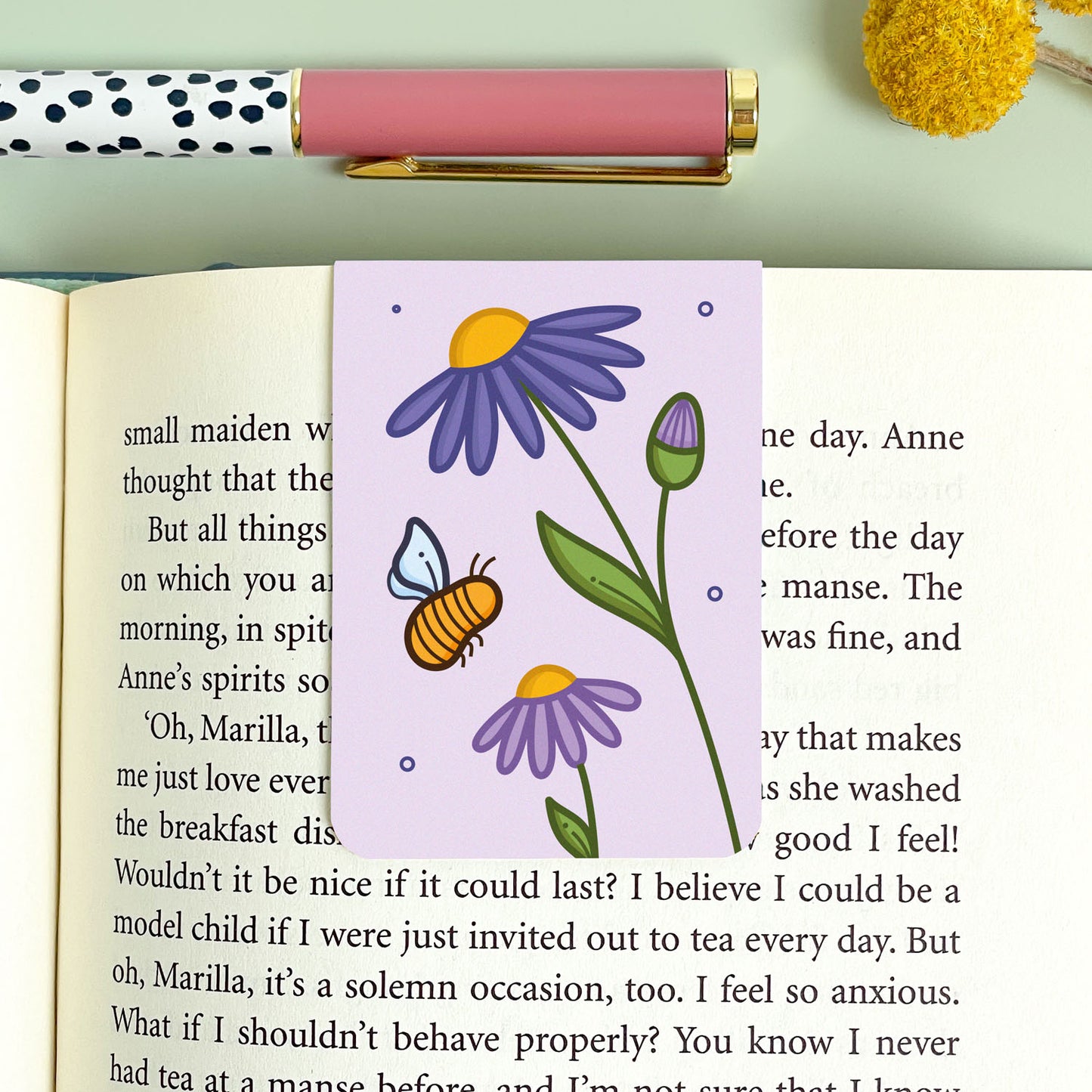 Book with a purple aster bookmark featuring a flower and bee design, placed on an open book.