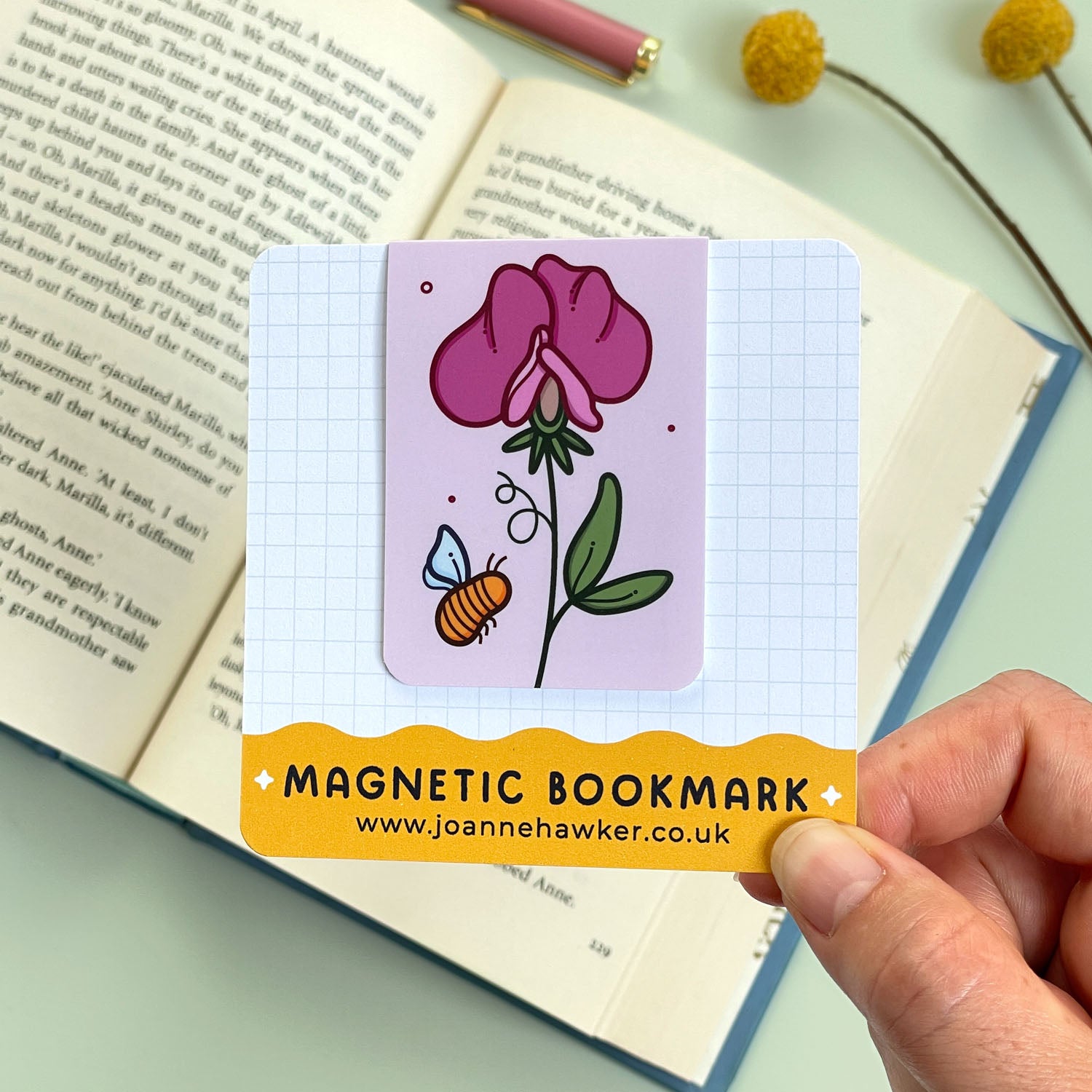 Magnetic bookmark on its backing card with pink floral sweet pea design held over an open book