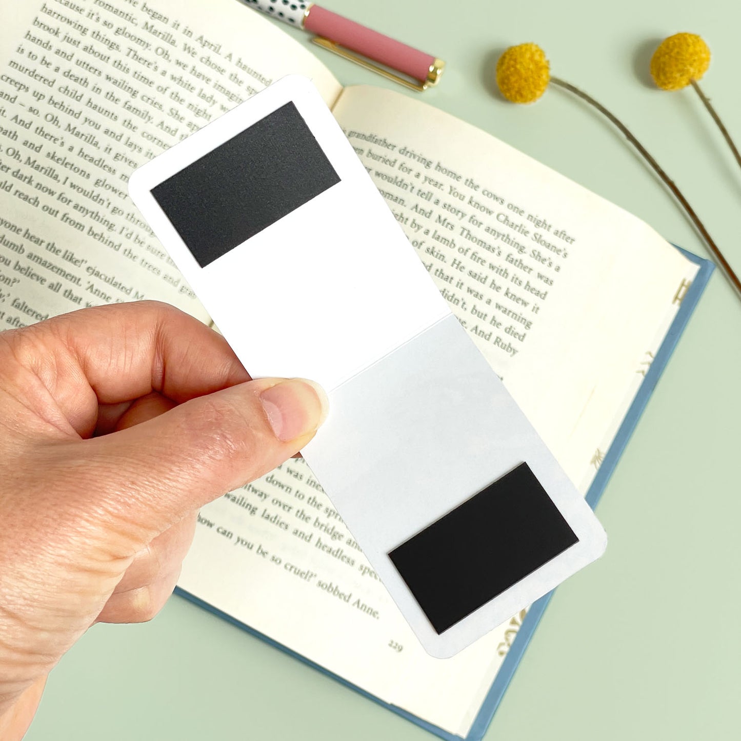 Hand holding an open bookmark to show the under side and the magnets over an open book with a pen and flowers on a light green surface.
