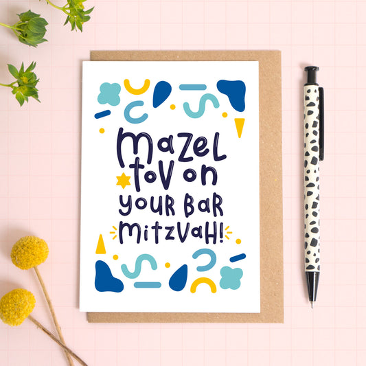 This Bar Mitzvah card has been photographed on top of a kraft brown envelope sitting upon a pink background with foliage and flowers to the left and a spotty pen to the right.