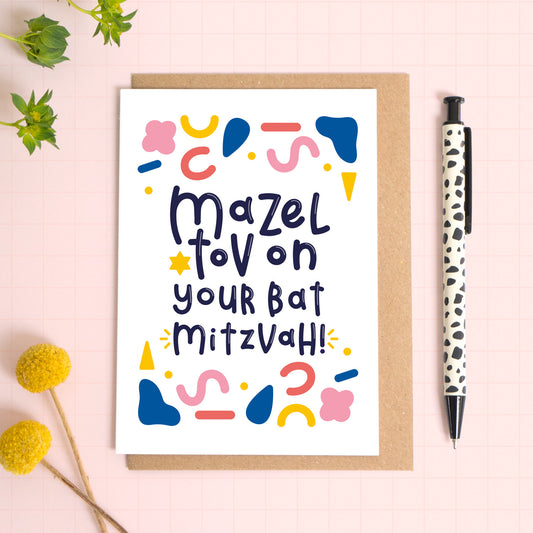 This Bat Mitzvah card has been photographed on top of a kraft brown envelope sitting upon a pink background with foliage and flowers to the left and a spotty pen to the right.