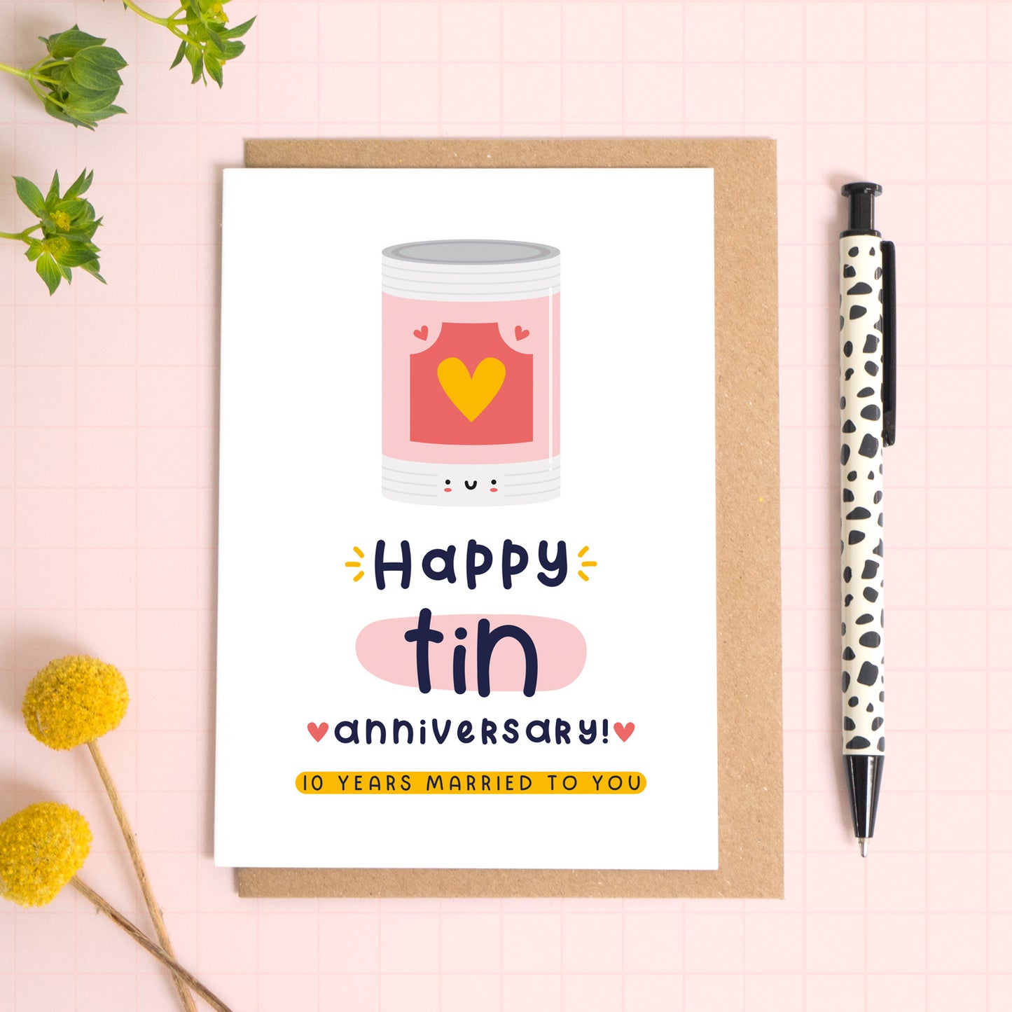 A ‘happy tin wedding anniversary’ card for a 10th wedding anniversary which has been photographed on a Kraft brown envelope upon a pink background surrounded by florals and a pen for scale.