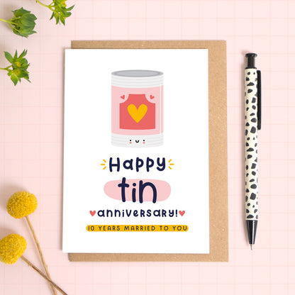 A ‘happy tin wedding anniversary’ card for a 10th wedding anniversary which has been photographed on a Kraft brown envelope upon a pink background surrounded by florals and a pen for scale.