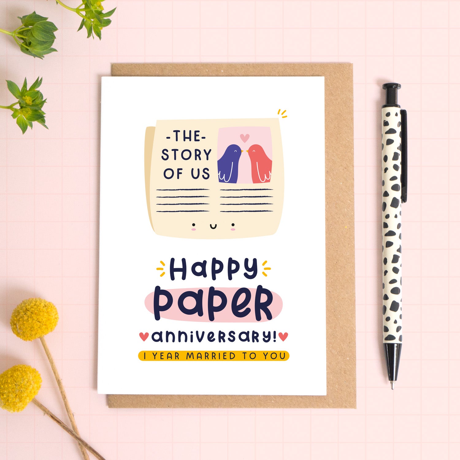 A ‘happy paper wedding anniversary’ card for a 1st wedding anniversary which has been photographed on a Kraft brown envelope upon a pink background surrounded by florals and a pen for scale.