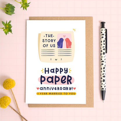 A ‘happy paper wedding anniversary’ card for a 1st wedding anniversary which has been photographed on a Kraft brown envelope upon a pink background surrounded by florals and a pen for scale.