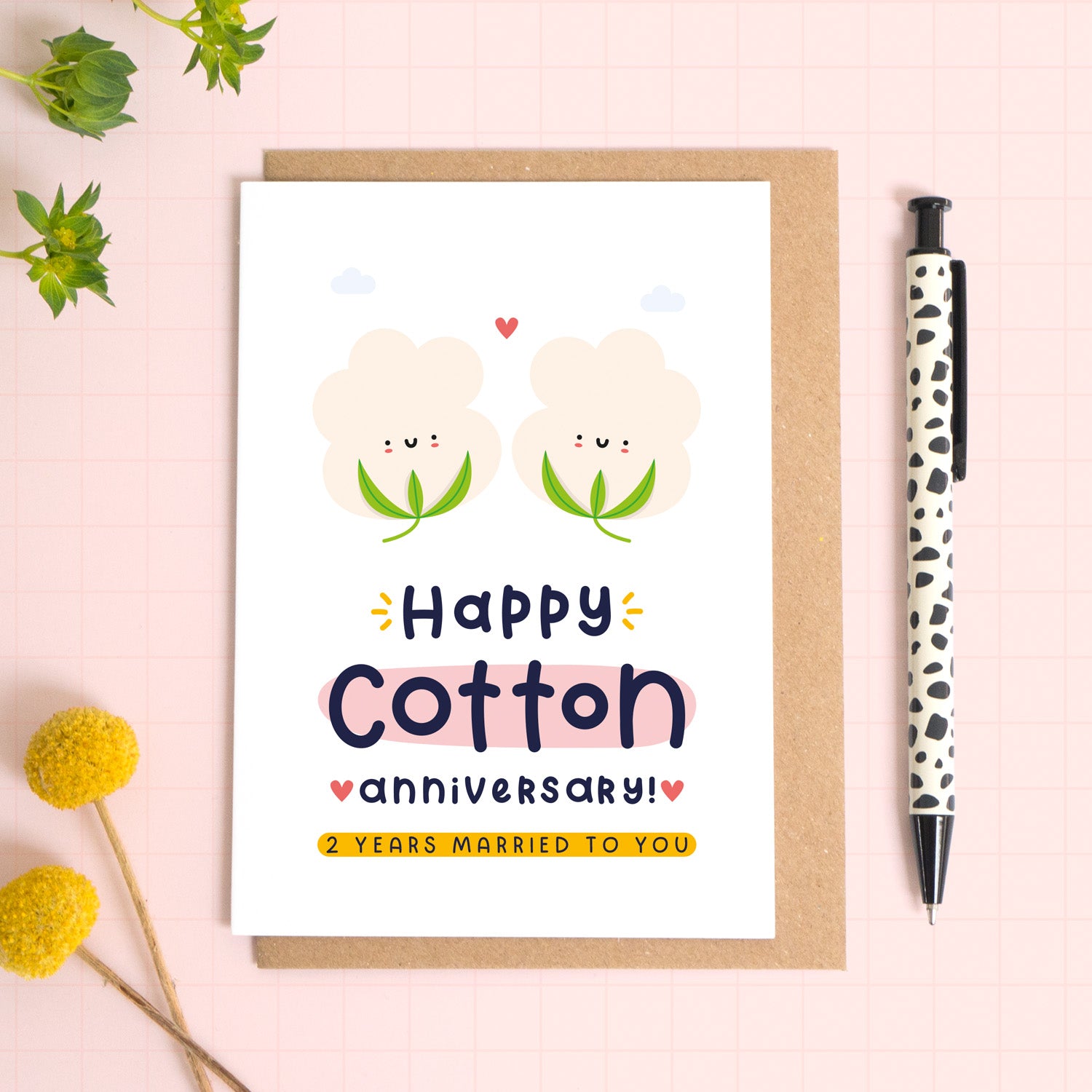 A ‘happy cotton wedding anniversary’ card for a 2nd wedding anniversary which has been photographed on a Kraft brown envelope upon a pink background surrounded by florals and a pen for scale.