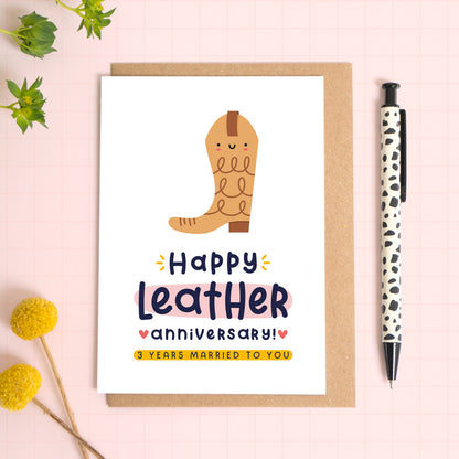 A ‘happy leather wedding anniversary’ card for a 3rd wedding anniversary which has been photographed on a Kraft brown envelope upon a pink background surrounded by florals and a pen for scale.