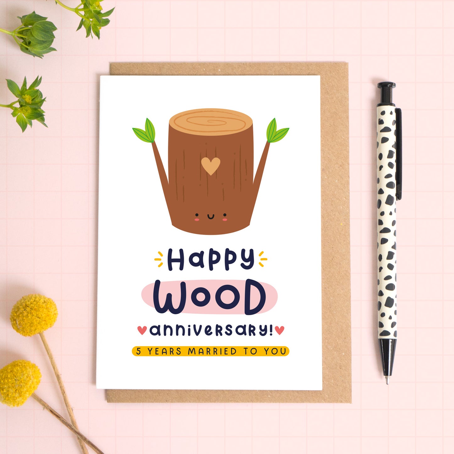 A ‘happy wood wedding anniversary’ card for a 5th wedding anniversary which has been photographed on a Kraft brown envelope upon a pink background surrounded by florals and a pen for scale.