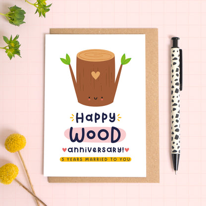 A ‘happy wood wedding anniversary’ card for a 5th wedding anniversary which has been photographed on a Kraft brown envelope upon a pink background surrounded by florals and a pen for scale.