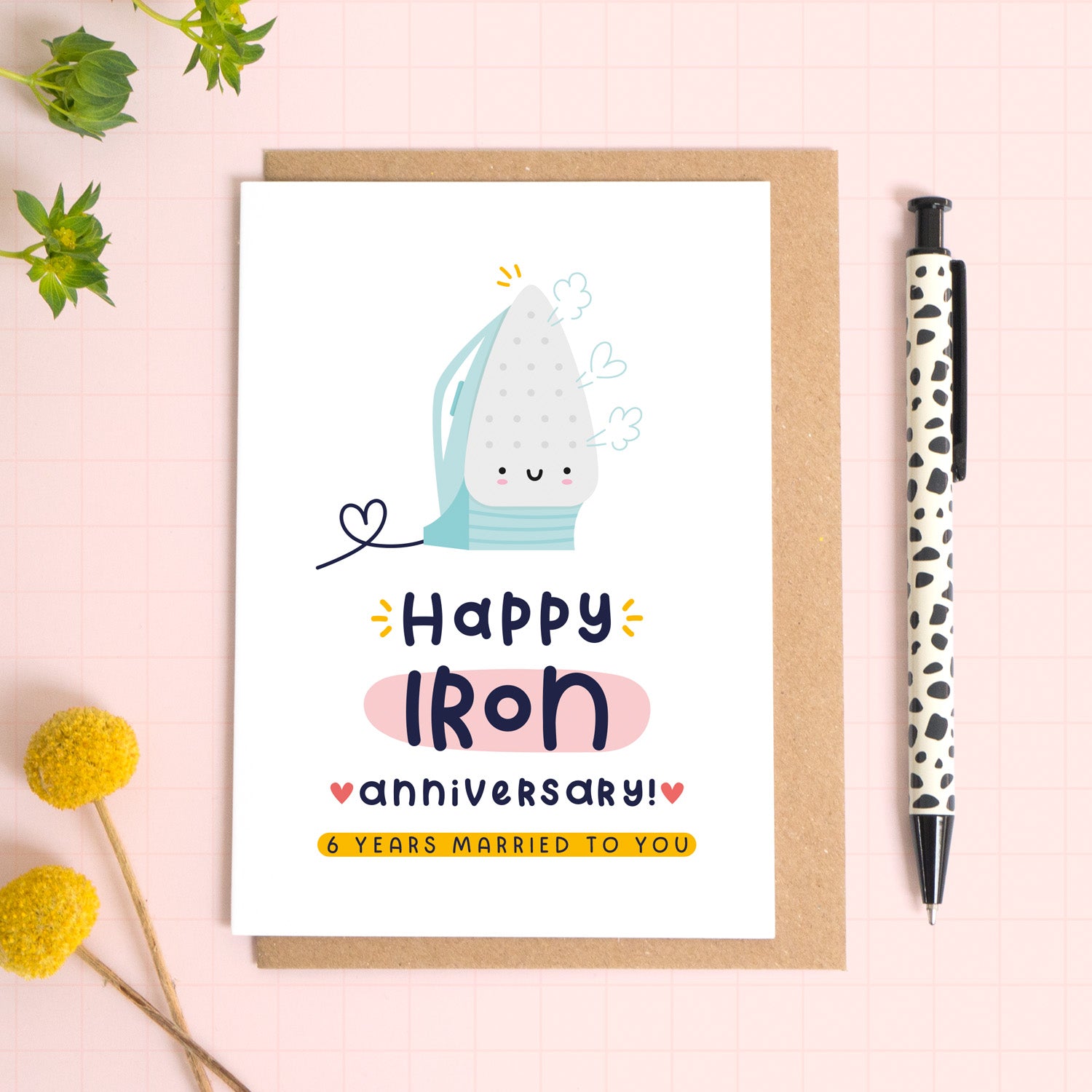 A ‘happy Iron wedding anniversary’ card for a 6th wedding anniversary which has been photographed on a Kraft brown envelope upon a pink background surrounded by florals and a pen for scale.