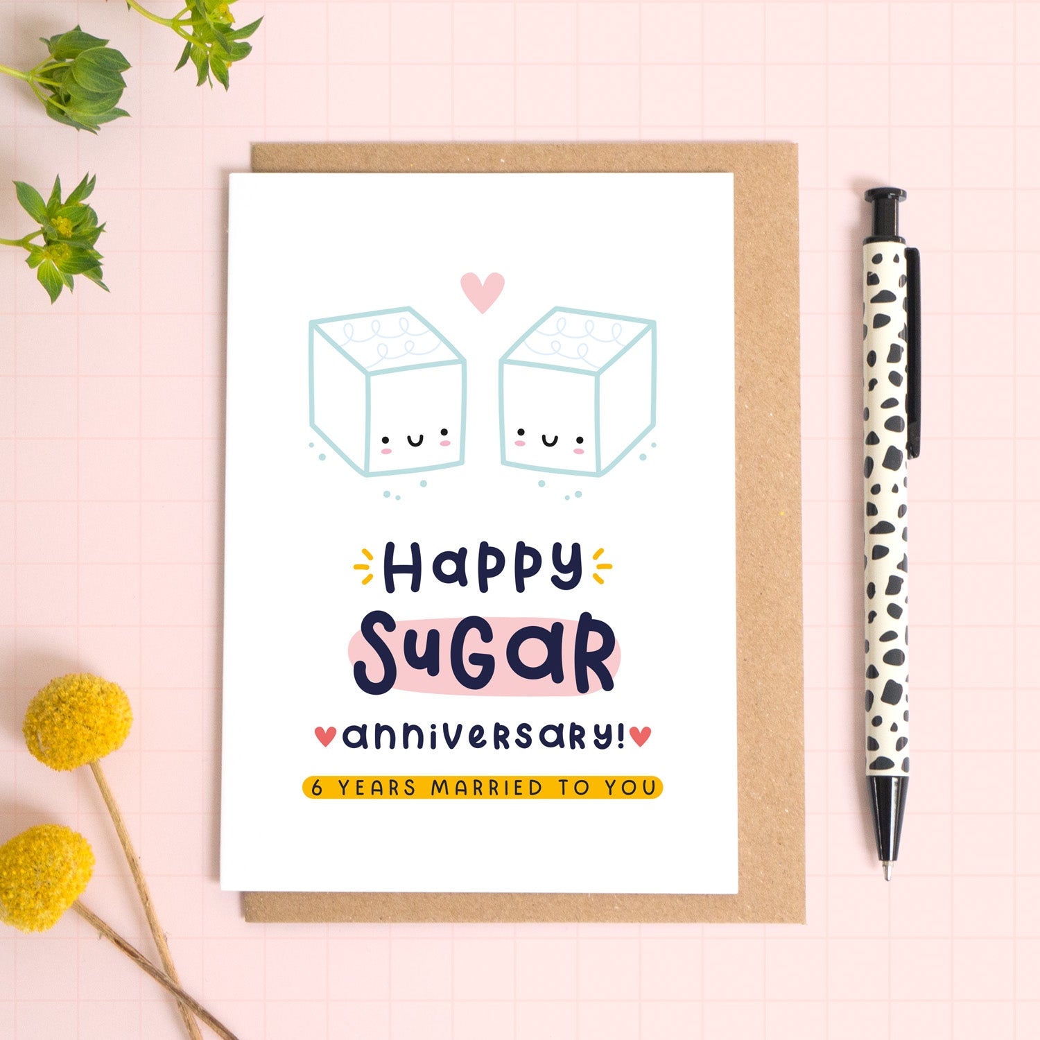 A ‘happy Sugar wedding anniversary’ card for a 6th wedding anniversary which has been photographed on a Kraft brown envelope upon a pink background surrounded by florals and a pen for scale.