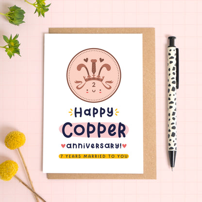 A ‘happy Copper wedding anniversary’ card for a 7th wedding anniversary which has been photographed on a Kraft brown envelope upon a pink background surrounded by florals and a pen for scale.