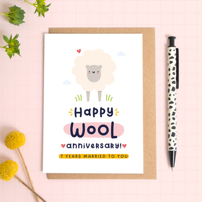 A ‘happy wool wedding anniversary’ card for a 7th wedding anniversary which has been photographed on a Kraft brown envelope upon a pink background surrounded by florals and a pen for scale.