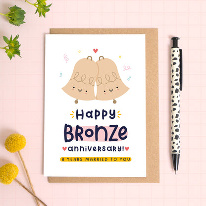 A ‘happy bronze wedding anniversary’ card for a 8th wedding anniversary which has been photographed on a Kraft brown envelope upon a pink background surrounded by florals and a pen for scale.