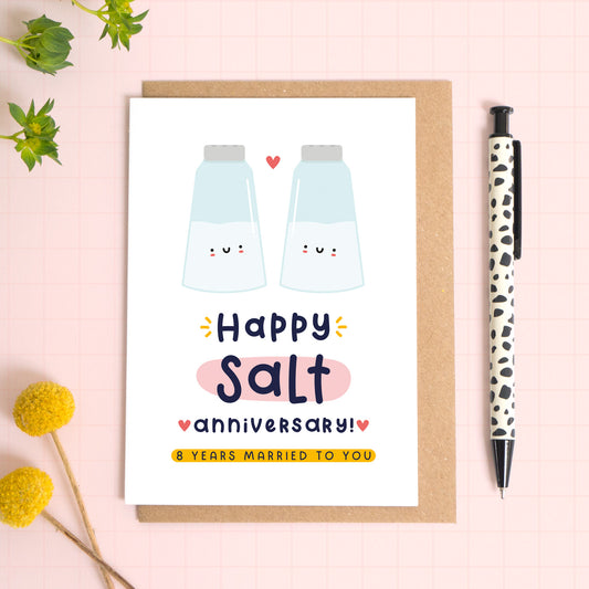 A ‘happy salt wedding anniversary’ card for a 8th wedding anniversary which has been photographed on a Kraft brown envelope upon a pink background surrounded by florals and a pen for scale.