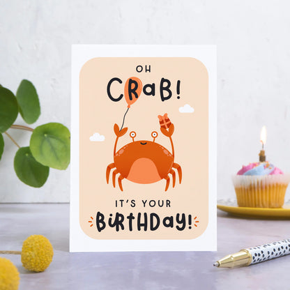 A birthday card featuring a crab holding a balloon and a present with the words ‘oh crab, it’s your birthday!’. The card is stood on a grey surface with foliage, flowers, a pen and a cupcake in the foreground and background.