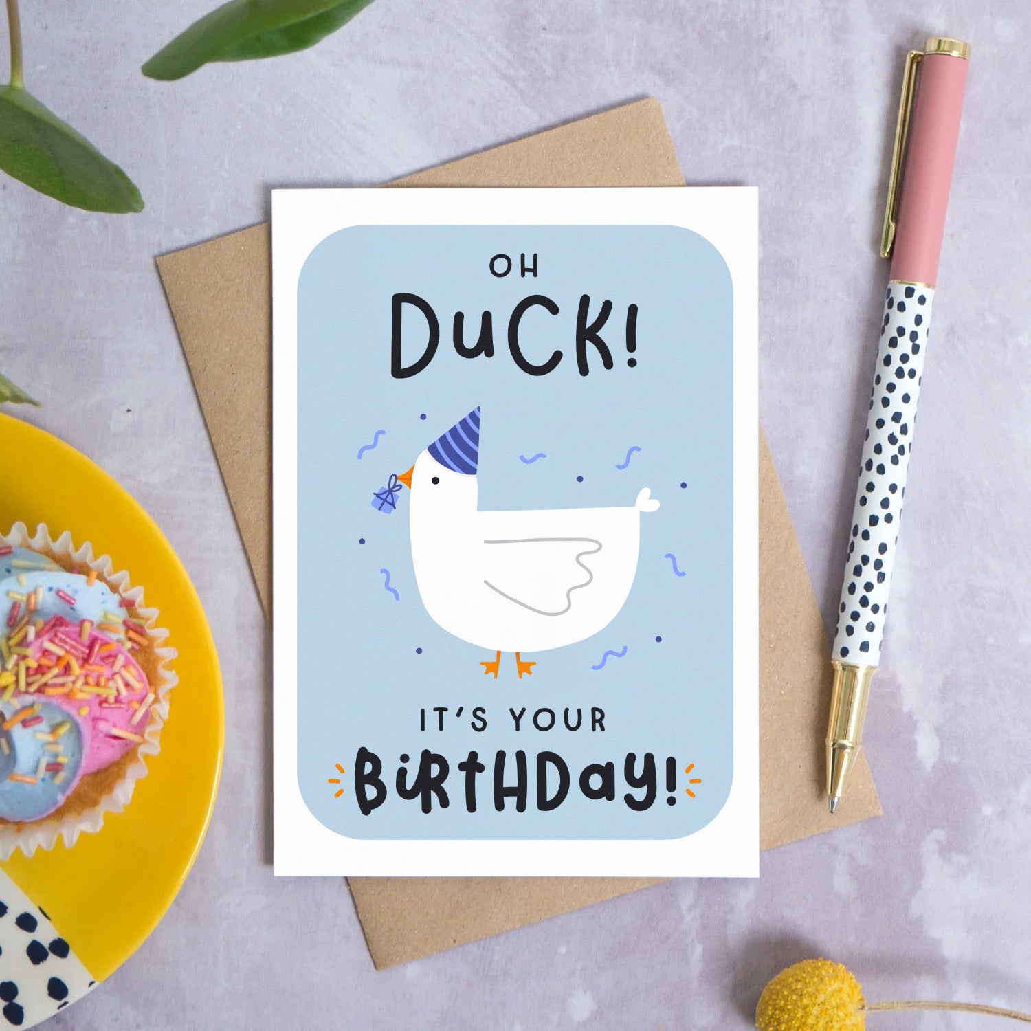 A birthday card featuring a duck in a party hat with a present in it’s beak with the words ‘oh duck, it’s your birthday!’. The card is lying on a Kraft brown envelope on a grey surface. Surrounding the card are leaves, flowers, a pen and a cupcake on a plate.