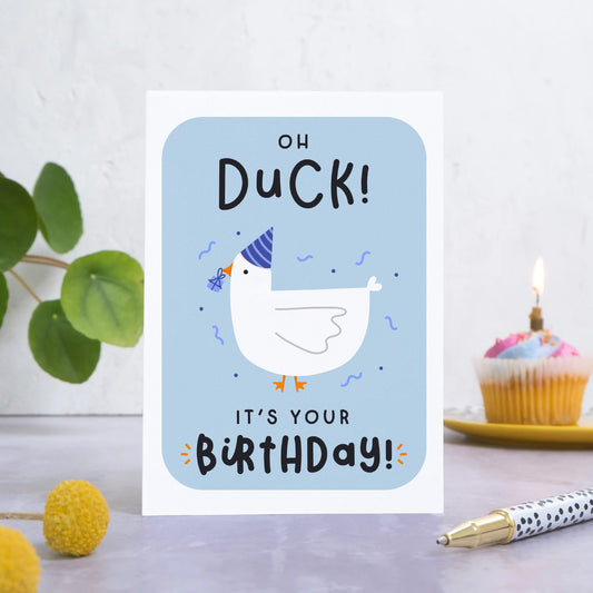 A birthday card featuring a duck in a party hat with a present in it’s beak with the words ‘oh duck, it’s your birthday!’. The card is stood on a grey surface with foliage, flowers, a pen and a cupcake in the foreground and background.