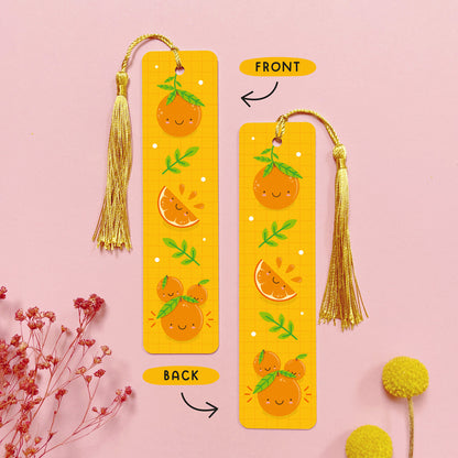 An image depicting the front and the back of the double sided metal bookmark which has been photographed on a pink background with dried flowers. Each bookmark has a gold tassel. The front and the back of the bookmark are printed with the same design of leaves and happy oranges.