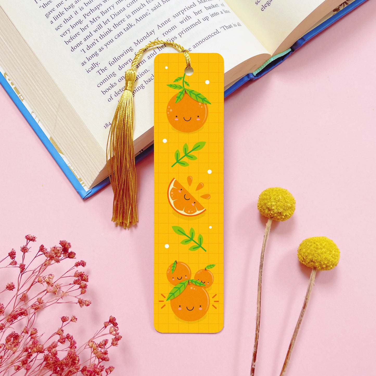 An orange themed, double sided, metal bookmark with a gold tassel which has been photographed on a pink background with an open book and dried flowers.