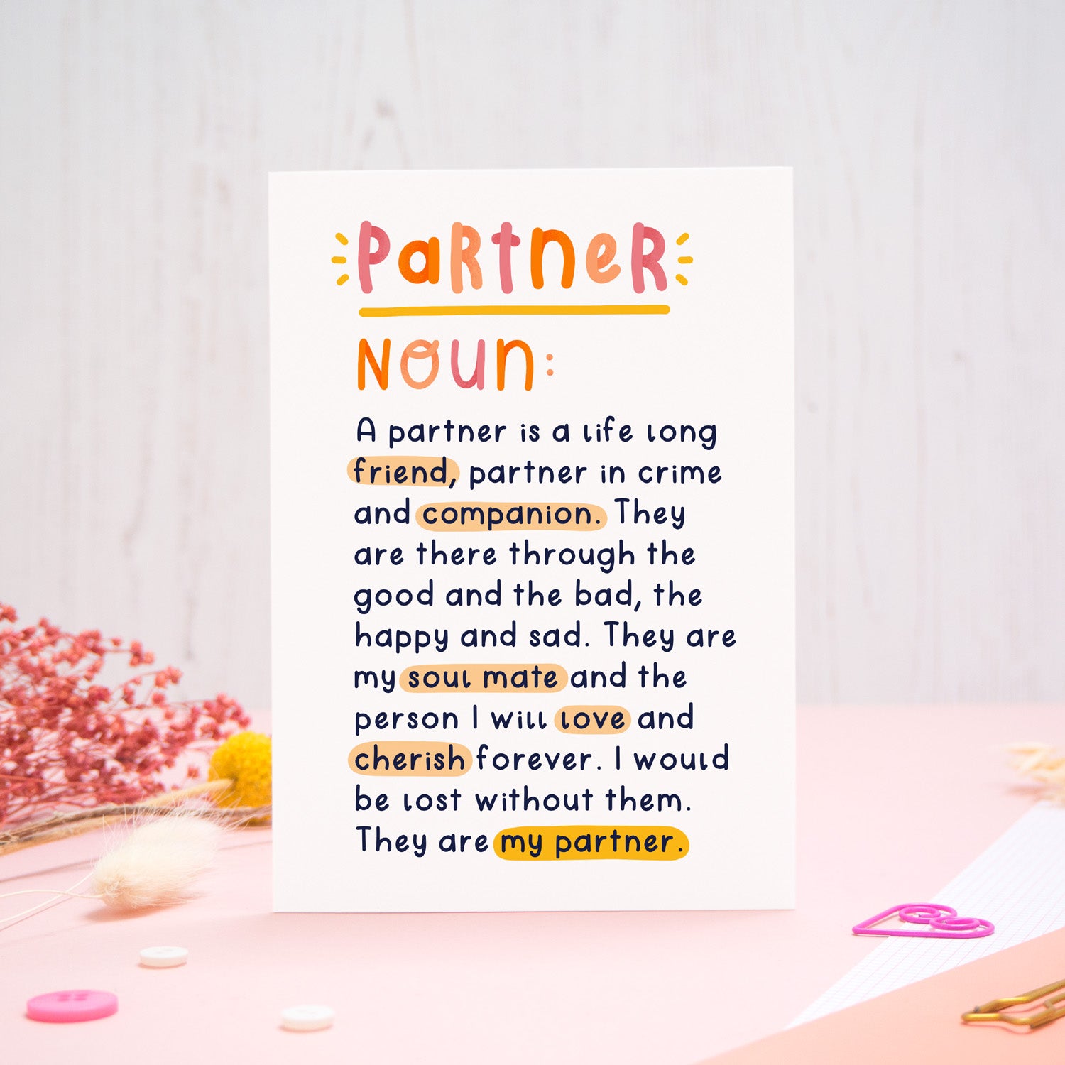A heartfelt, colourful dictionary definition style card with the definition of a partner, photographed stood on a pink and white background with floral props, paper clips, and buttons. 
