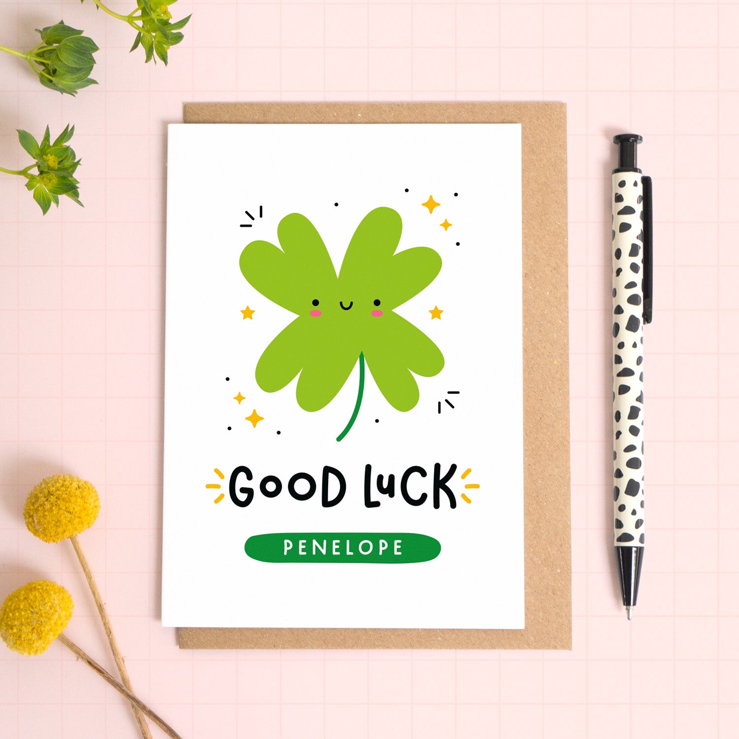 A personalised good luck card with an illustration of a four leaf clover, and the recipients name printed on the front of the card. The card has been photographed on it's kraft brown envelope on a pink surface with foliage and a pen for scale.