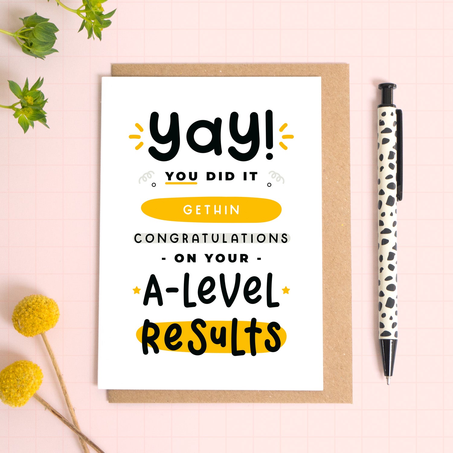 A personalised A-Level exam results congratulations card for someone who has just received brilliant exam results. This card has been photographed on top of the Kraft brown envelope that it comes with and on top of a pink background. There is foliage to the left and a pen on the right for scale. This is the black and yellow version.