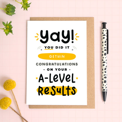 A personalised A-Level exam results congratulations card for someone who has just received brilliant exam results. This card has been photographed on top of the Kraft brown envelope that it comes with and on top of a pink background. There is foliage to the left and a pen on the right for scale. This is the black and yellow version.