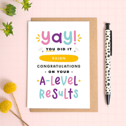 A personalised A-Level exam results congratulations card for someone who has just received brilliant exam results. This card has been photographed on top of the Kraft brown envelope that it comes with and on top of a pink background. There is foliage to the left and a pen on the right for scale. This is the pink, purple and blue version.