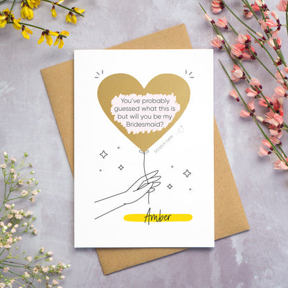 The personalised question scratch card has been photographed lying on top of a Kraft brown envelope which is on top of a grey surface surrounded by colourful flowers. The gold heart scratch panel has been scratched away to reveal the surprise question.