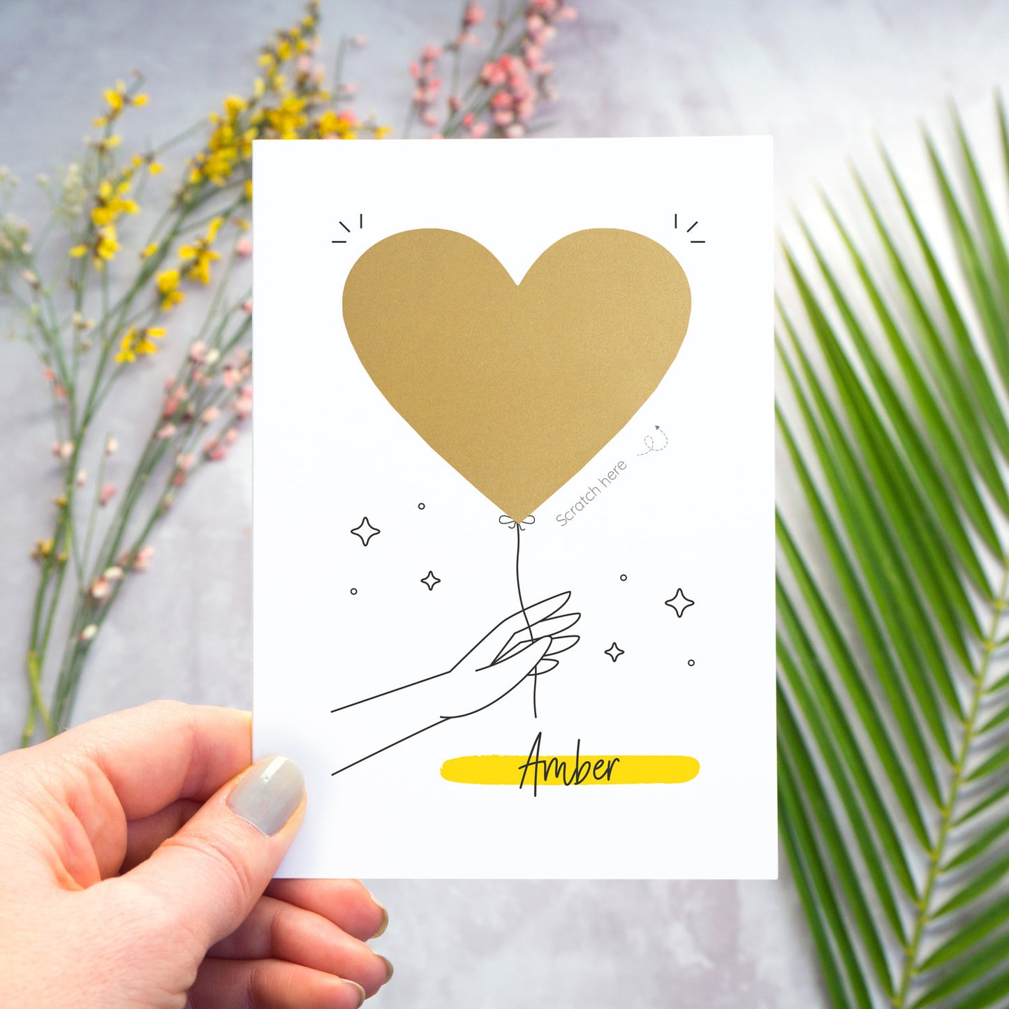 This image shows how the personalised question scratch card looks before the recipient scratches away the heart. In this image the card is being held over a grey surface which has a variety of colour flowers and foliage in the background. The heart has not yet been scratched away.