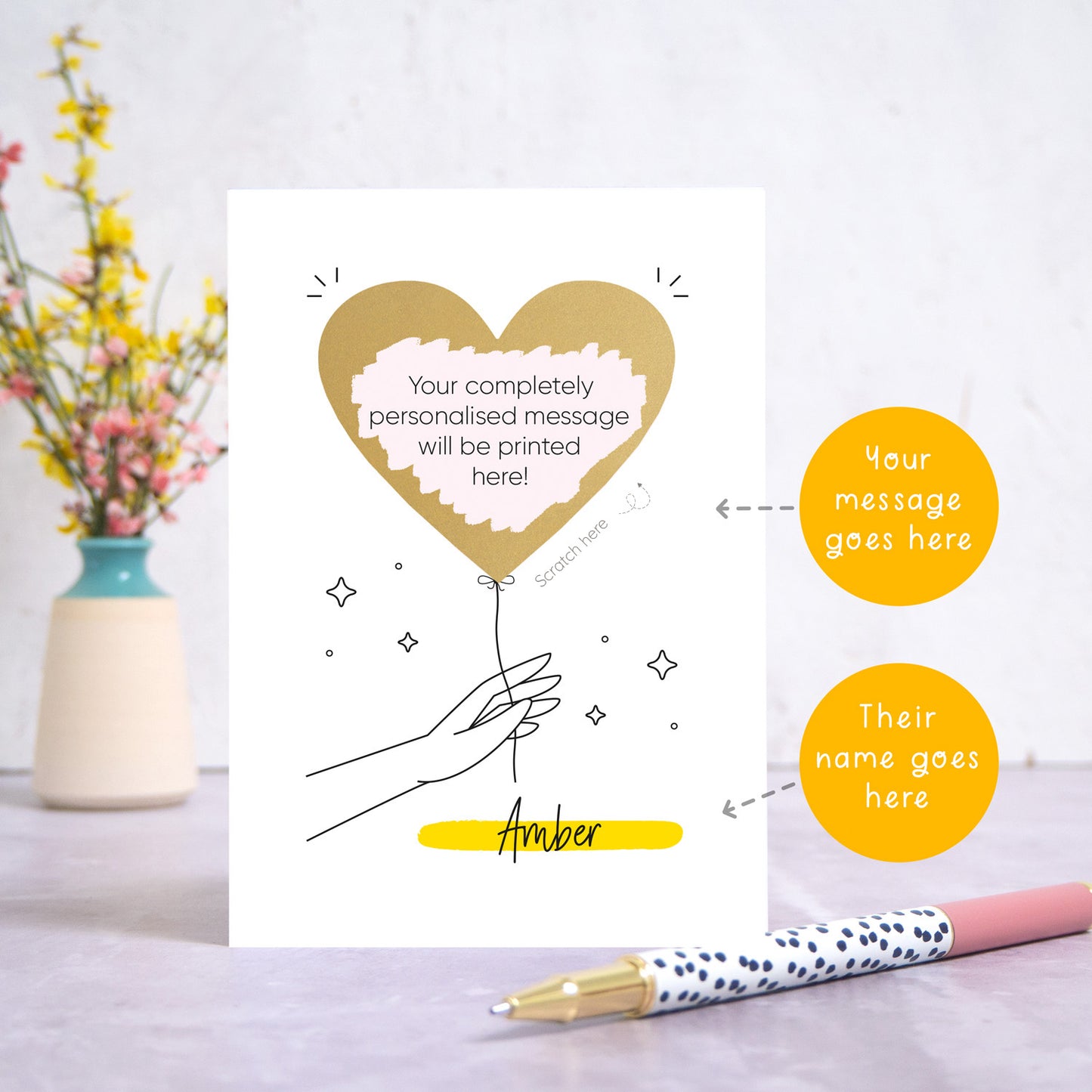 The scratch and reveal card has been photographed standing up on top of a grey surface. There is a small jar of flowers in the background and a pen in the foreground. To the right of the card are two circles which point to the areas that can be personalised. These are the name and the scratch off message. The gold heart has been scratched off to reveal the custom text.