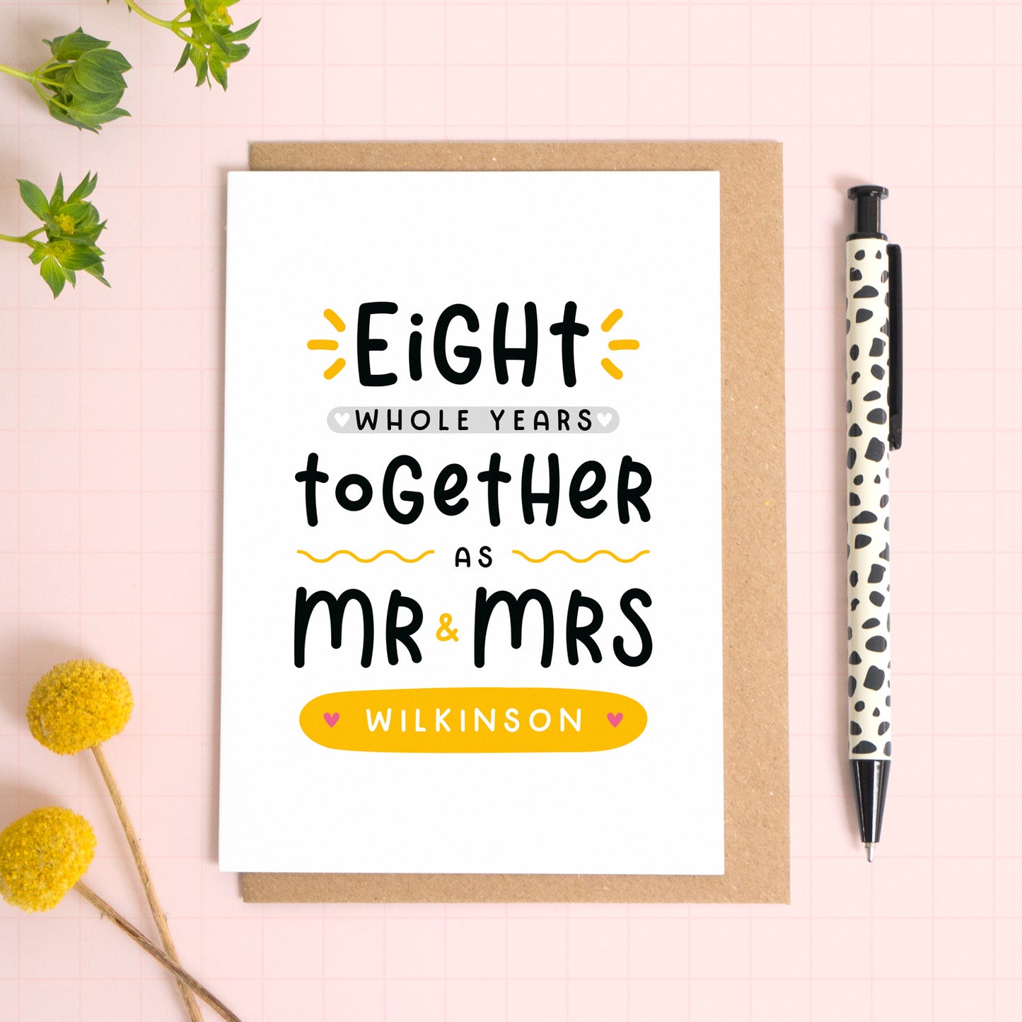 A personalised Mr & Mrs anniversary card reading ‘eight whole years together as Mr & Mrs [surname]’ which has been photographed on a Kraft brown envelope upon a pink background surrounded by florals and a pen for scale.