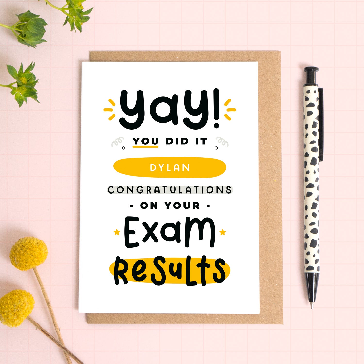 A personalised exam results congratulations card for someone who has just received brilliant exam results. This card has been photographed on top of the Kraft brown envelope that it comes with and on top of a pink background. There is foliage to the left and a pen on the right for scale. This is the black and yellow version.