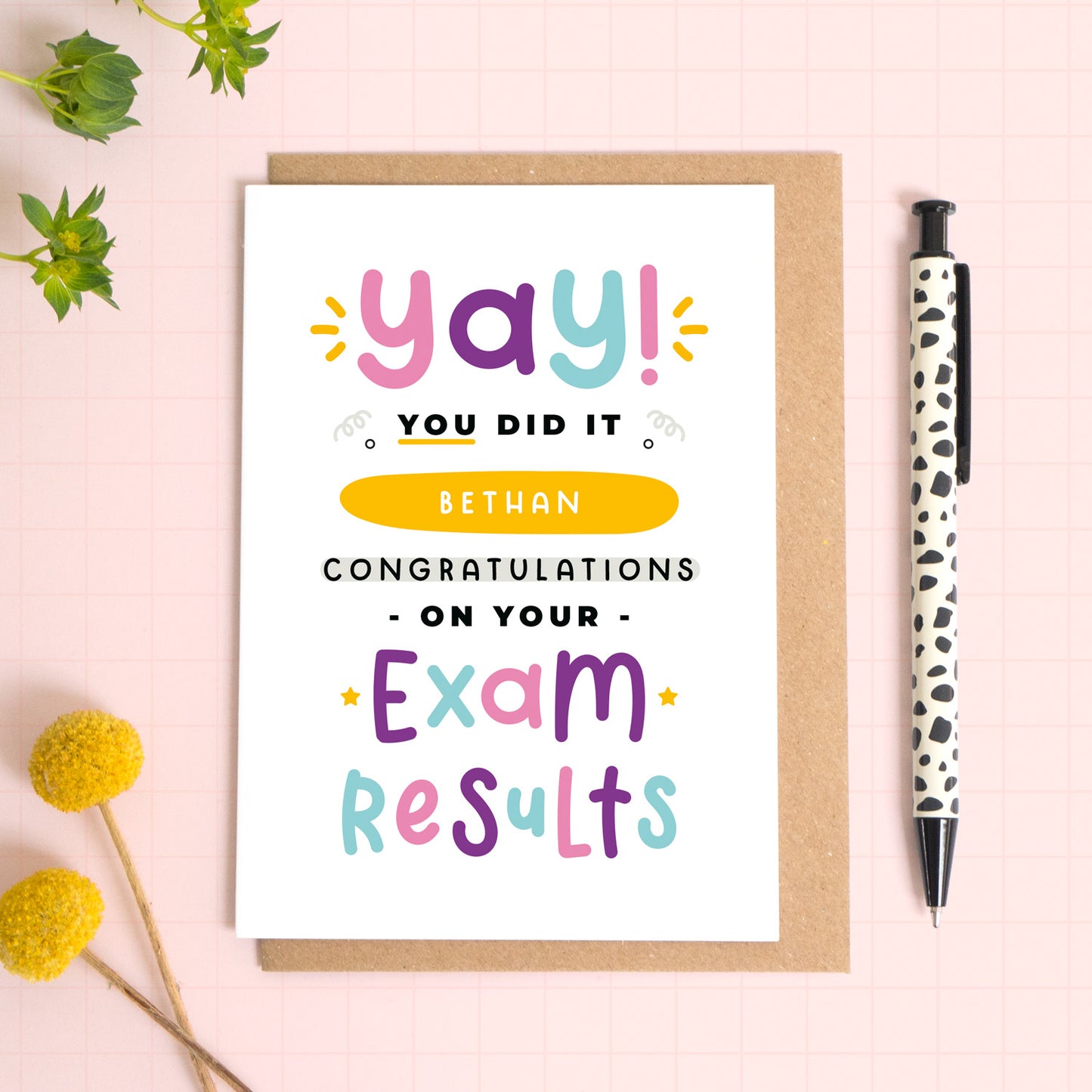 A personalised exam results congratulations card for someone who has just received brilliant exam results. This card has been photographed on top of the Kraft brown envelope that it comes with and on top of a pink background. There is foliage to the left and a pen on the right for scale. This is the pink, purple and blue version.