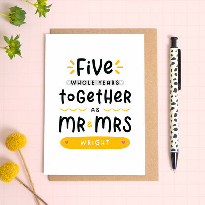 A personalised Mr & Mrs anniversary card reading ‘five whole years together as Mr & Mrs [surname]’ which has been photographed on a Kraft brown envelope upon a pink background surrounded by florals and a pen for scale.