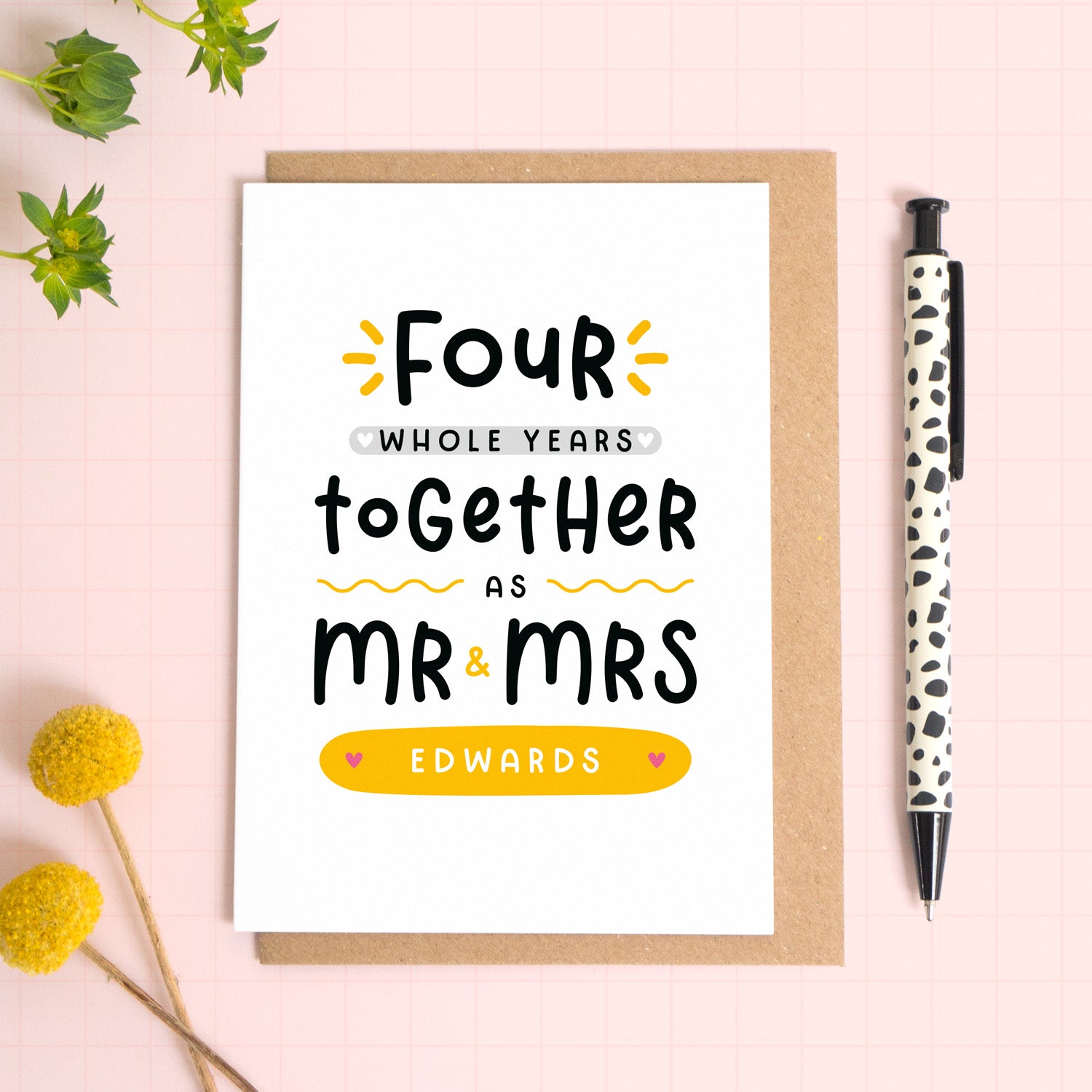 A personalised Mr & Mrs anniversary card reading ‘four whole years together as Mr & Mrs [surname]’ which has been photographed on a Kraft brown envelope upon a pink background surrounded by florals and a pen for scale.