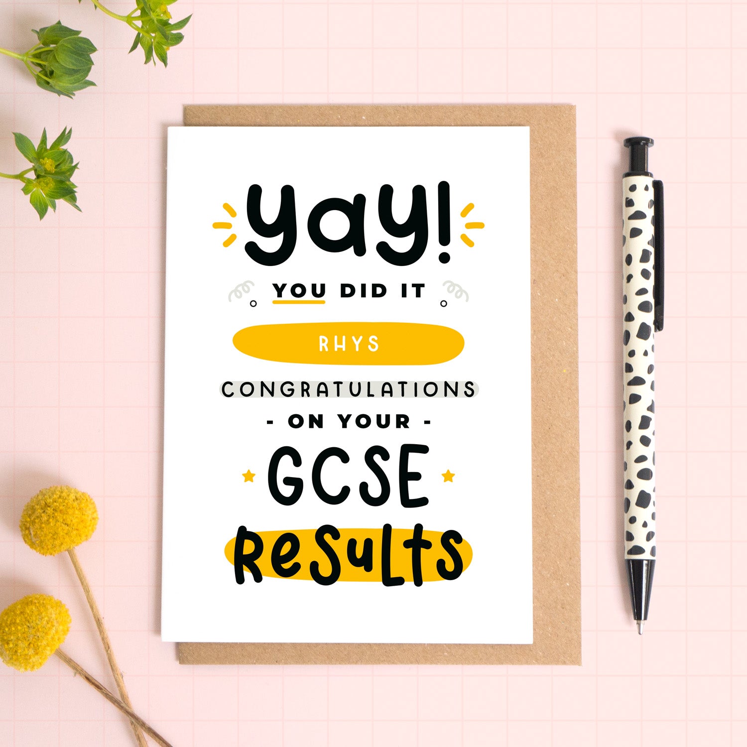 A personalised GCSE exam results congratulations card for someone who has just received brilliant exam results. This card has been photographed on top of the Kraft brown envelope that it comes with and on top of a pink background. There is foliage to the left and a pen on the right for scale. This is the black and yellow version.