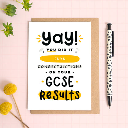 A personalised GCSE exam results congratulations card for someone who has just received brilliant exam results. This card has been photographed on top of the Kraft brown envelope that it comes with and on top of a pink background. There is foliage to the left and a pen on the right for scale. This is the black and yellow version.