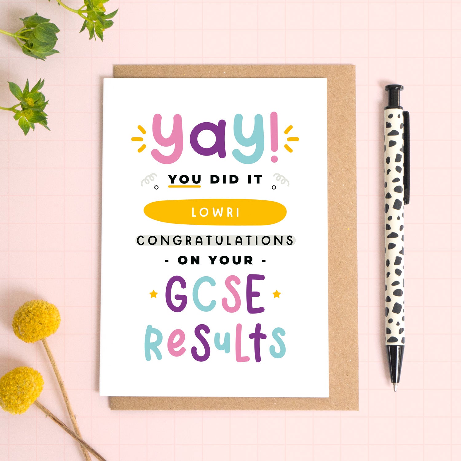 A personalised GCSE exam results congratulations card for someone who has just received brilliant exam results. This card has been photographed on top of the Kraft brown envelope that it comes with and on top of a pink background. There is foliage to the left and a pen on the right for scale. This is the pink, purple and blue version.