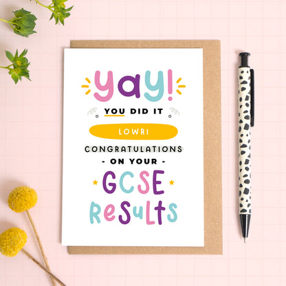 A personalised GCSE exam results congratulations card for someone who has just received brilliant exam results. This card has been photographed on top of the Kraft brown envelope that it comes with and on top of a pink background. There is foliage to the left and a pen on the right for scale. This is the pink, purple and blue version.