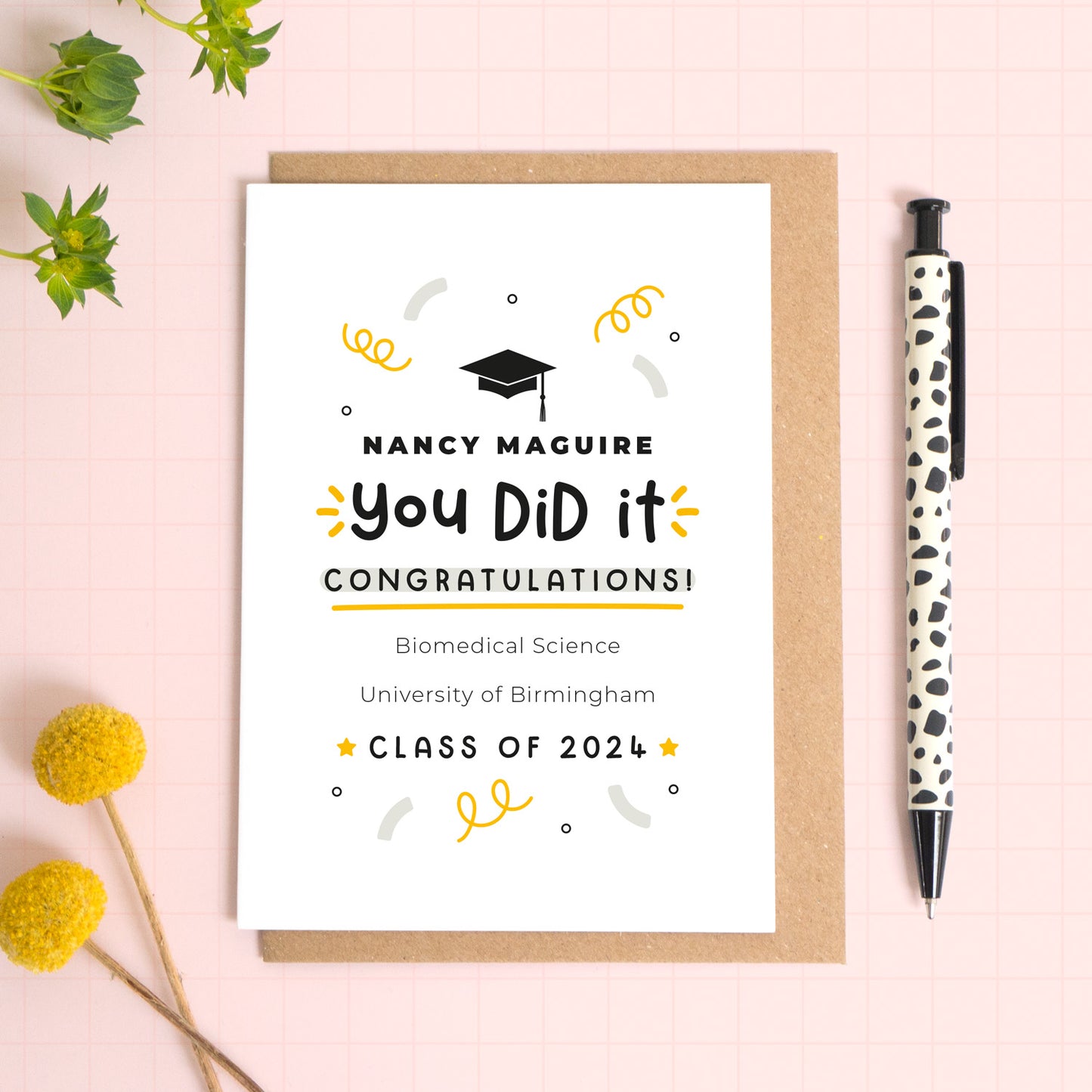 A personalised graduation card featuring a graduation cap, the recipients name, course title, university and class year and confetti lying down on top of a kraft brown envelope. To the side is foliage and a pen for scale. 