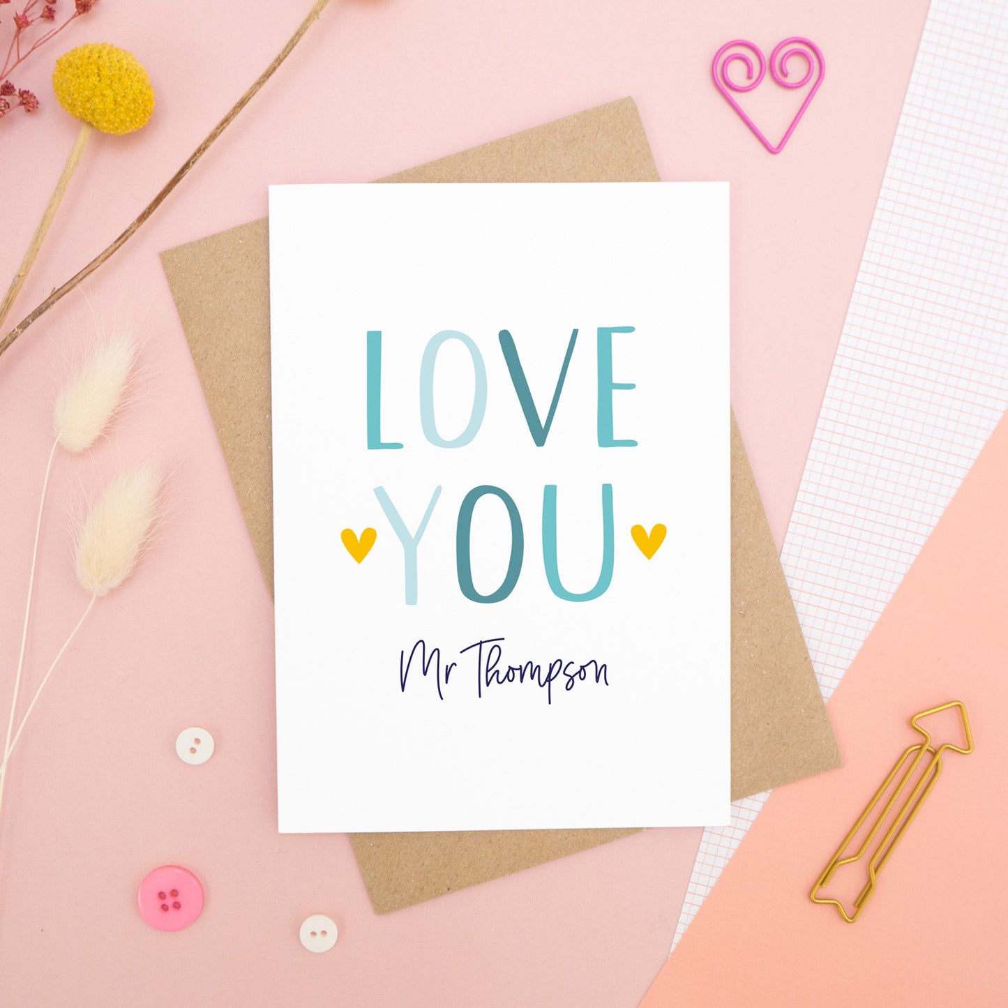 A personalised love you card photographed on a pink background with floral props, paper clips, and buttons. This image shows the ‘love you’ anniversary card in the blue colour way. The personalisation is navy and there are pops of yellow in the hearts.