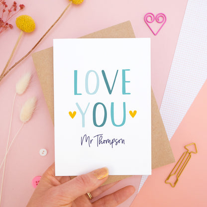 A personalised love you card photographed held over on a pink background with floral props, paper clips, and buttons. This image shows the ‘love you’ anniversary card in the blue colour way. The personalisation is navy and there are pops of yellow in the hearts.