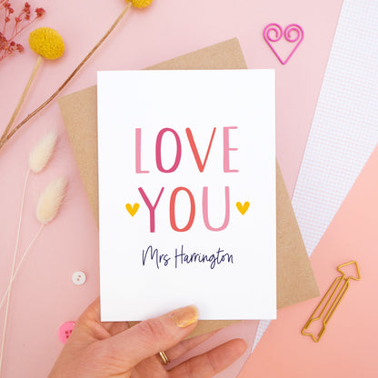 A personalised love you card photographed held over on a pink background with floral props, paper clips, and buttons. This image shows the ‘love you’ anniversary card in the pink colour way. The personalisation is navy and there are pops of yellow in the hearts.