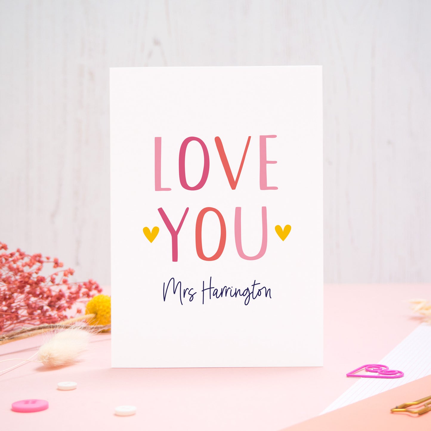 A personalised love you card photographed stood on a pink and white background with floral props, paper clips, and buttons. This image shows the ‘love you’ anniversary card in the pink colour way. The personalisation is navy and there are pops of yellow in the hearts.