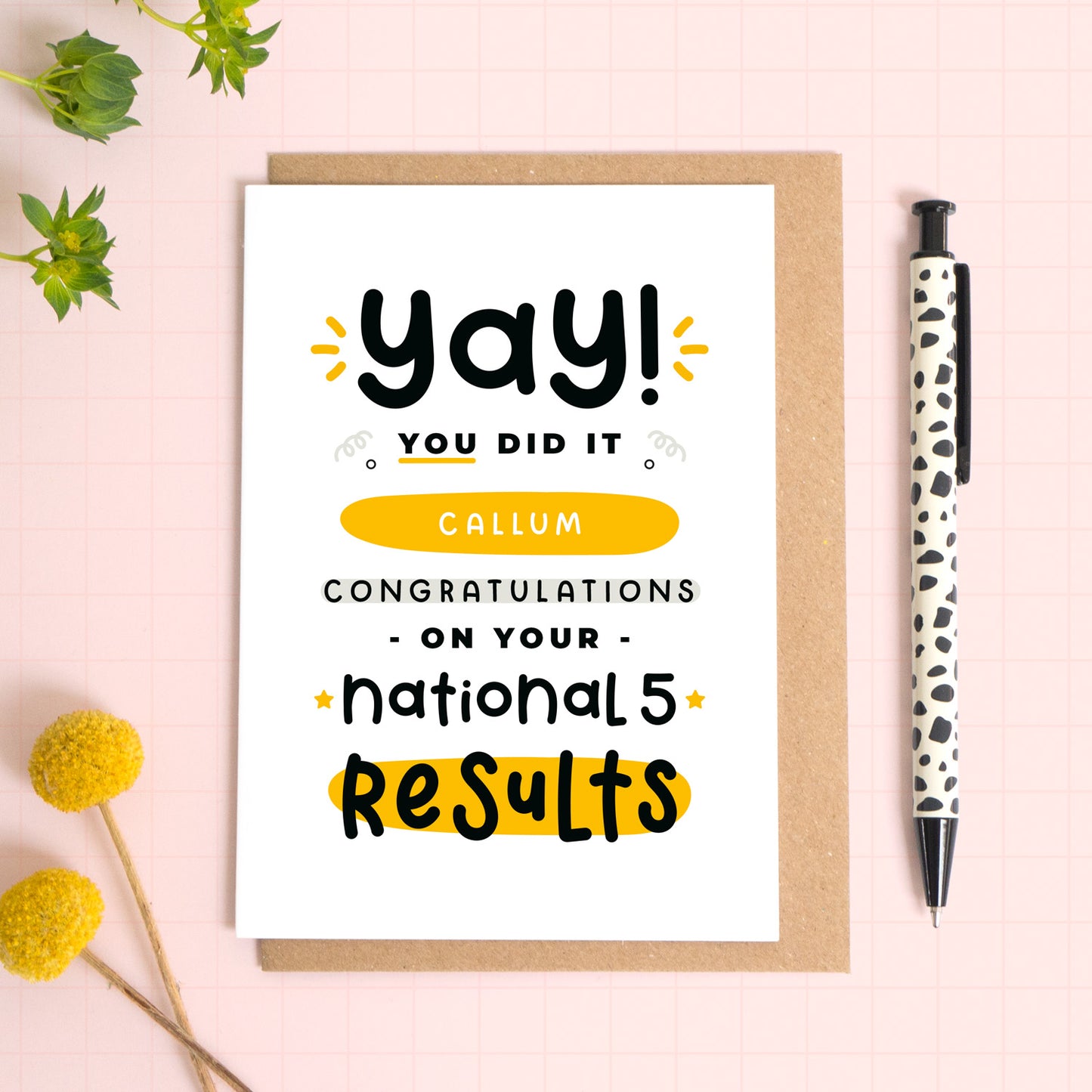 A personalised National 5 exam results congratulations card for someone who has just received brilliant exam results. This card has been photographed on top of the Kraft brown envelope that it comes with and on top of a pink background. There is foliage to the left and a pen on the right for scale. This is the black and yellow version.