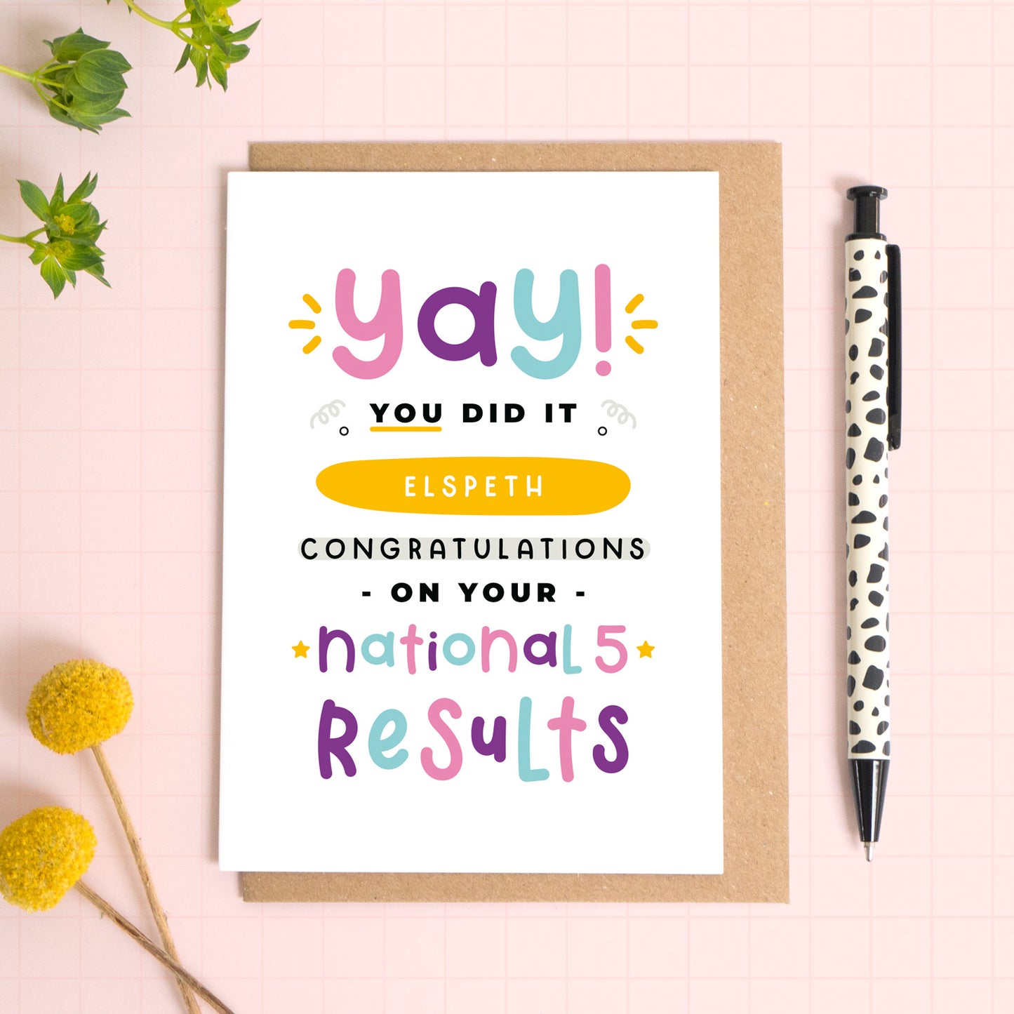 A personalised National 5 exam results congratulations card for someone who has just received brilliant exam results. This card has been photographed on top of the Kraft brown envelope that it comes with and on top of a pink background. There is foliage to the left and a pen on the right for scale. This is the pink, purple and blue version.